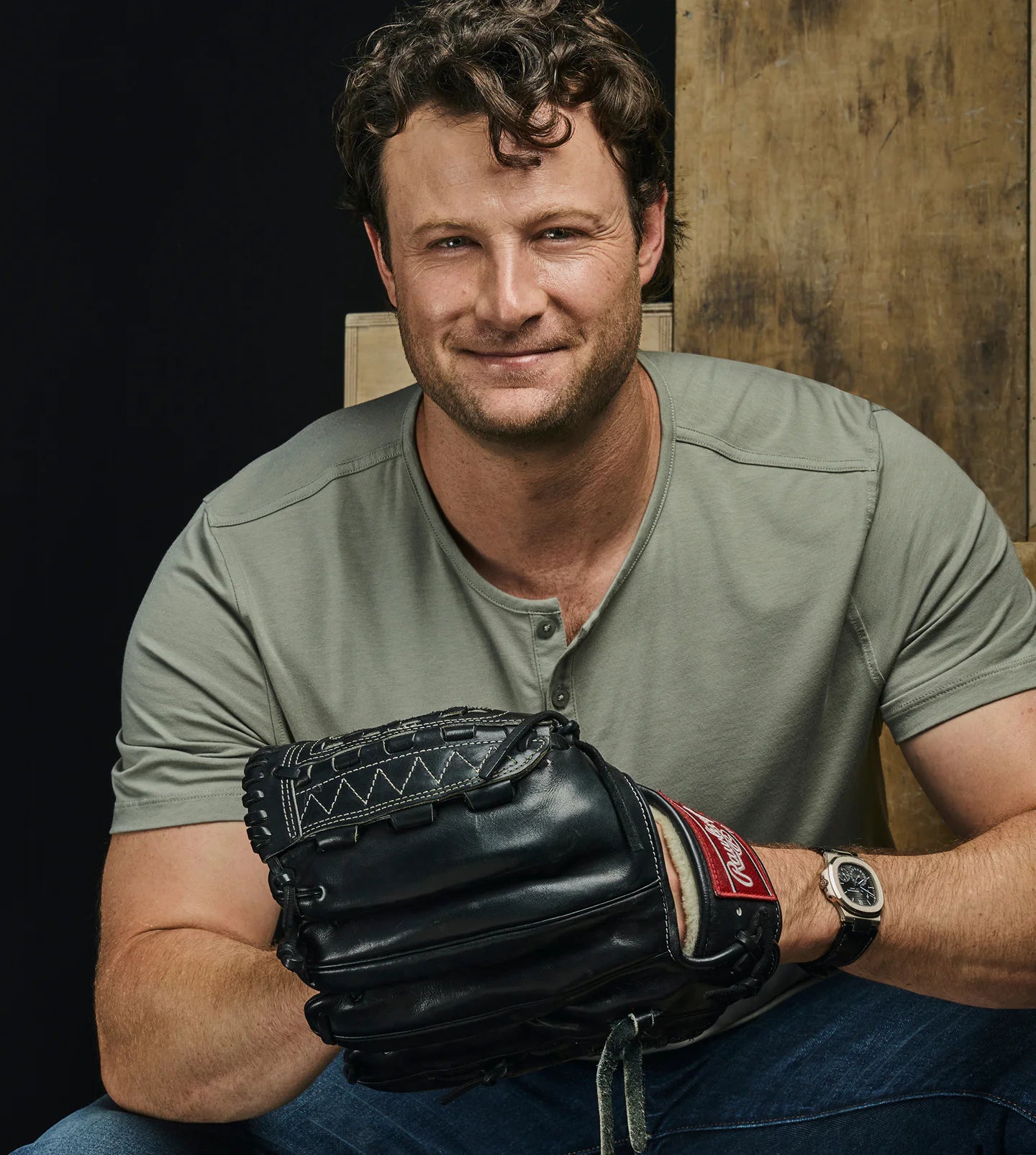 Man holding a baseball glove, sitting on wooden blocks, with a serious expression.